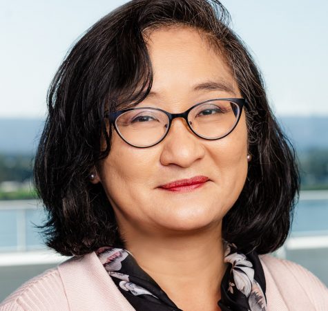 A woman with short dark hair wearing a pink sweater, a black neck scarf, and glasses smiles for the camera.
