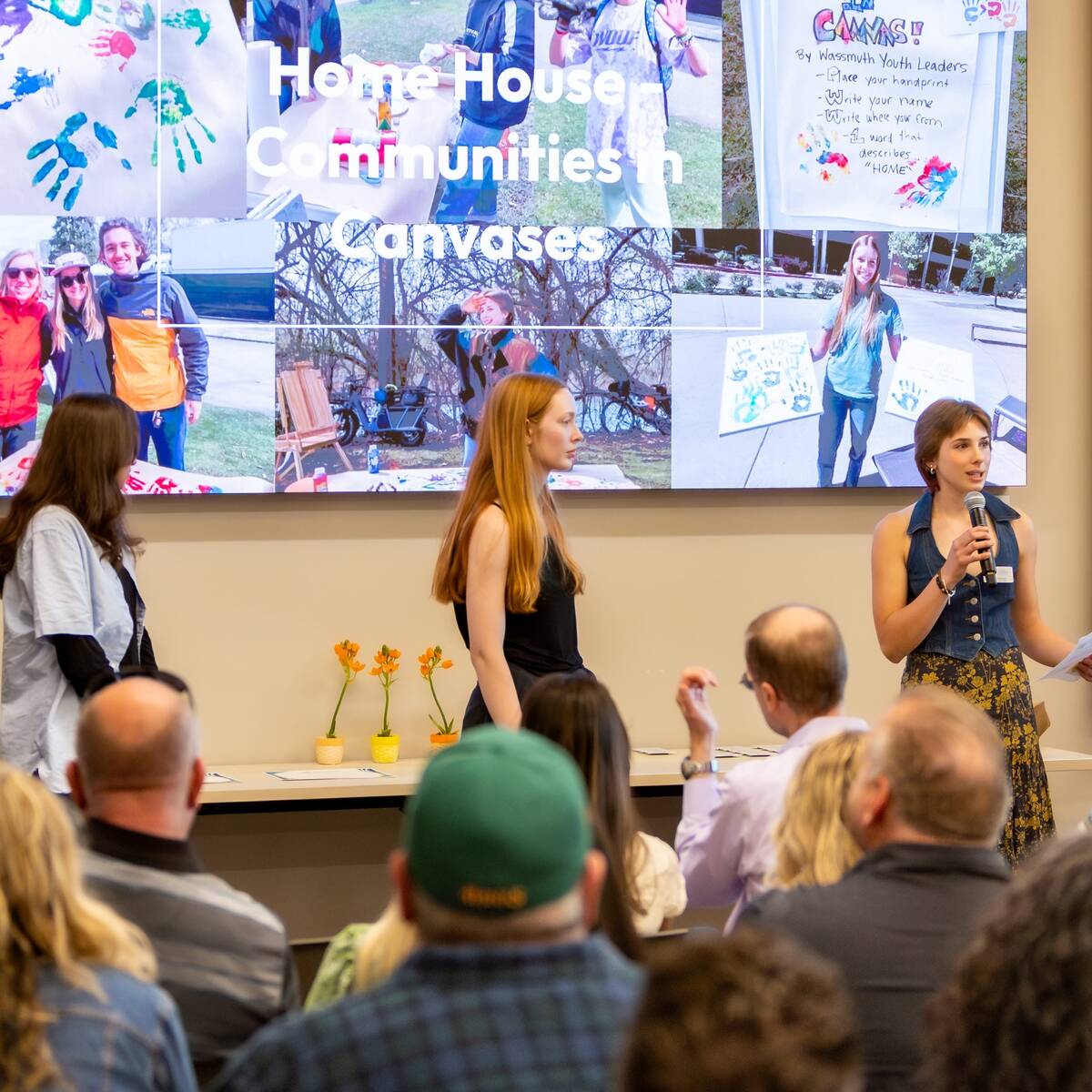 Five people standing at the front of a room of seated individuals. One person speaking into a microphone, holding a piece of paper.