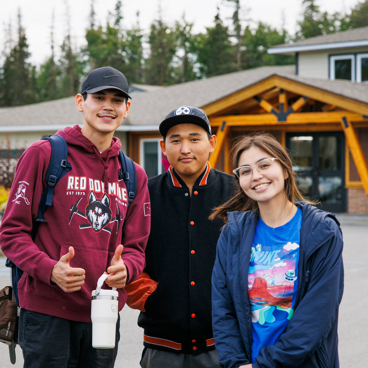 Three students posing and smiling outside in front of a building. One person holding a water bottle and two thumbs up.