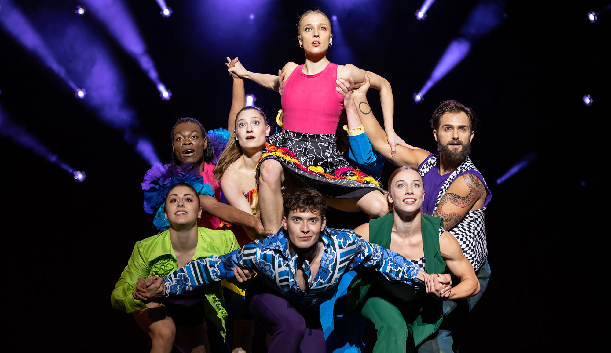 Seven dancers on stage in the middle of a performance with dramatic lighting behind them