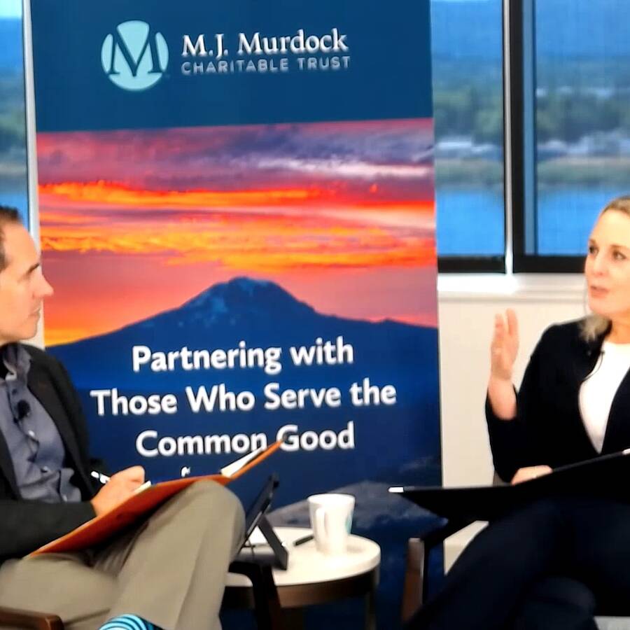Two people sitting in chairs in front of a Murdock Trust branded sign. One person speaking and the other intently listening.