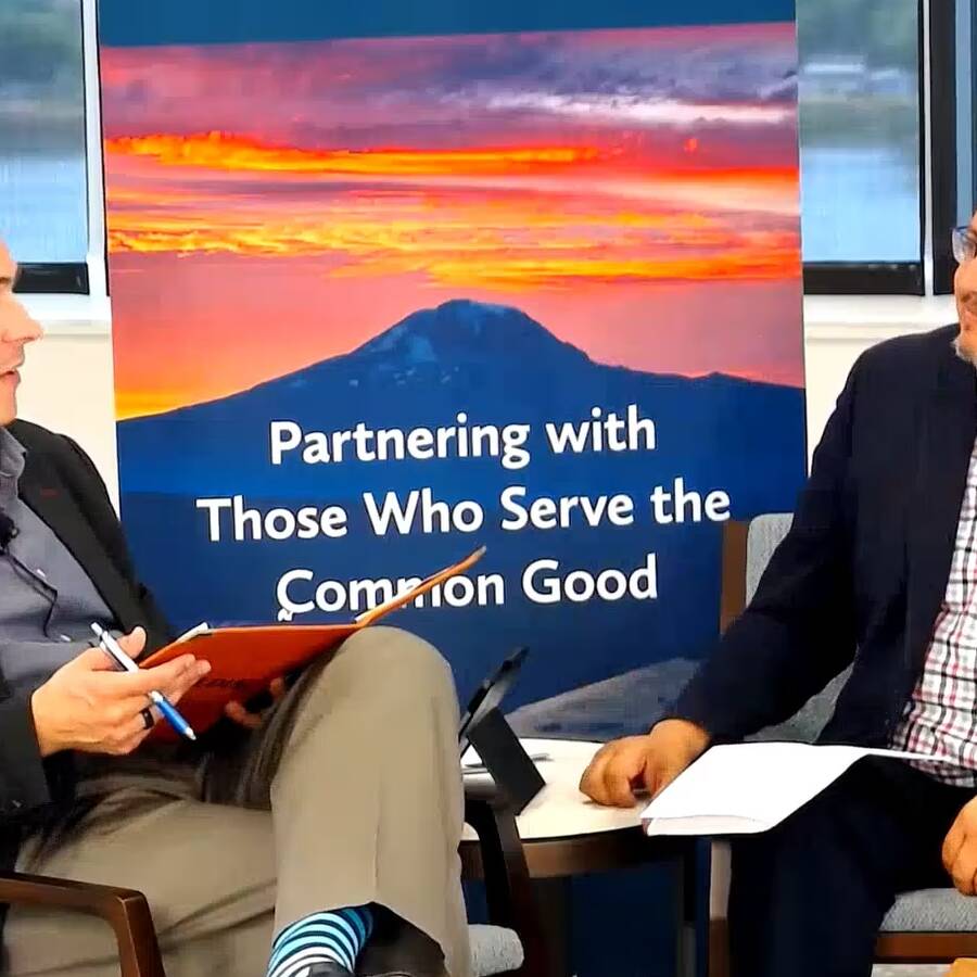 Two people sitting in front of a Murdock Trust branded sign. Both are in professional attire, speaking to each other.