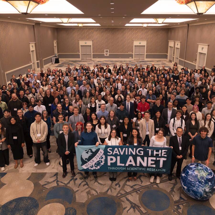 Group of people standing in conference room. People in the front holding up a sign that says, 'Saving the Planet Through Scientific Research.'