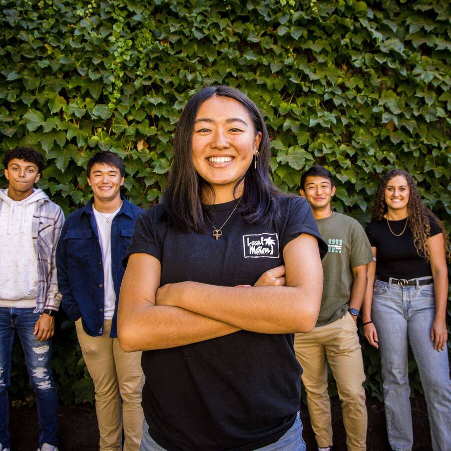 One student standing in the foreground with arms crossed and a big smile. Six additional students smiling and standing in the background against a wall covered in green leaves.