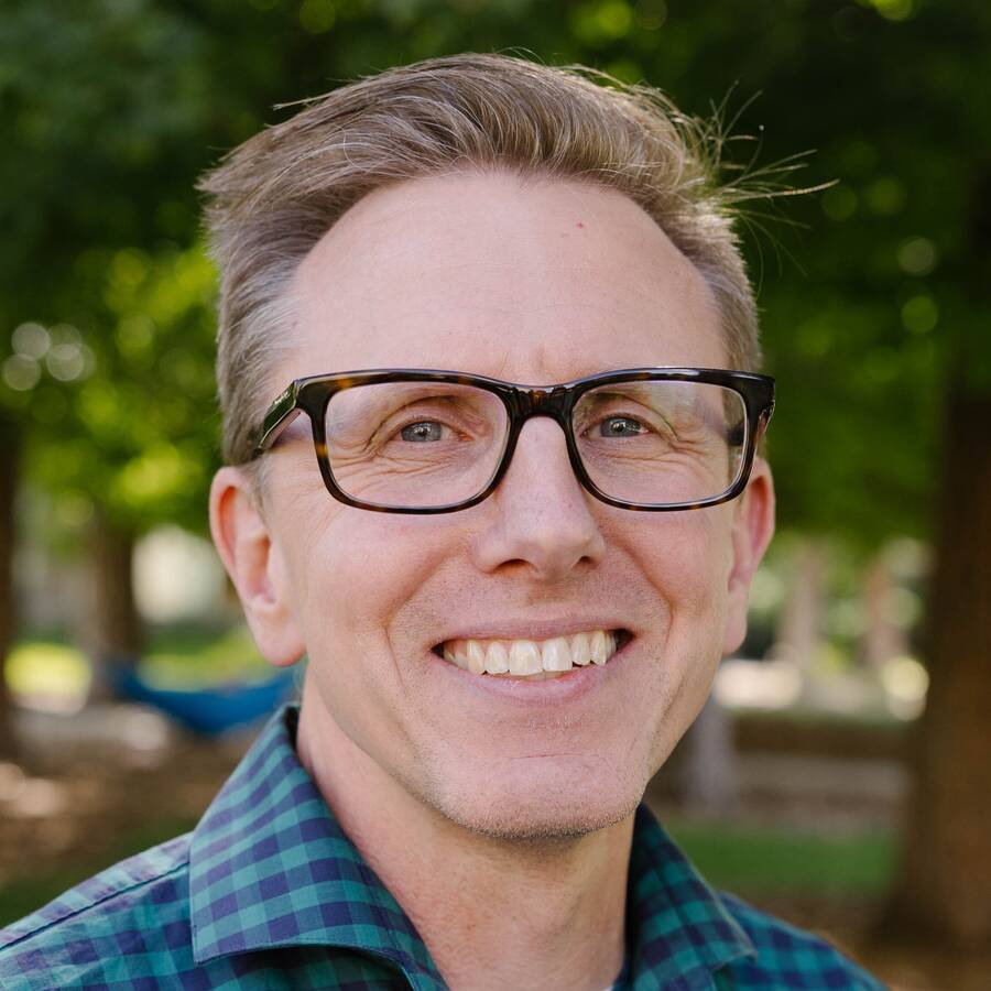 Headshot of person with glasses, smiling, and outside somewhere sunny with trees