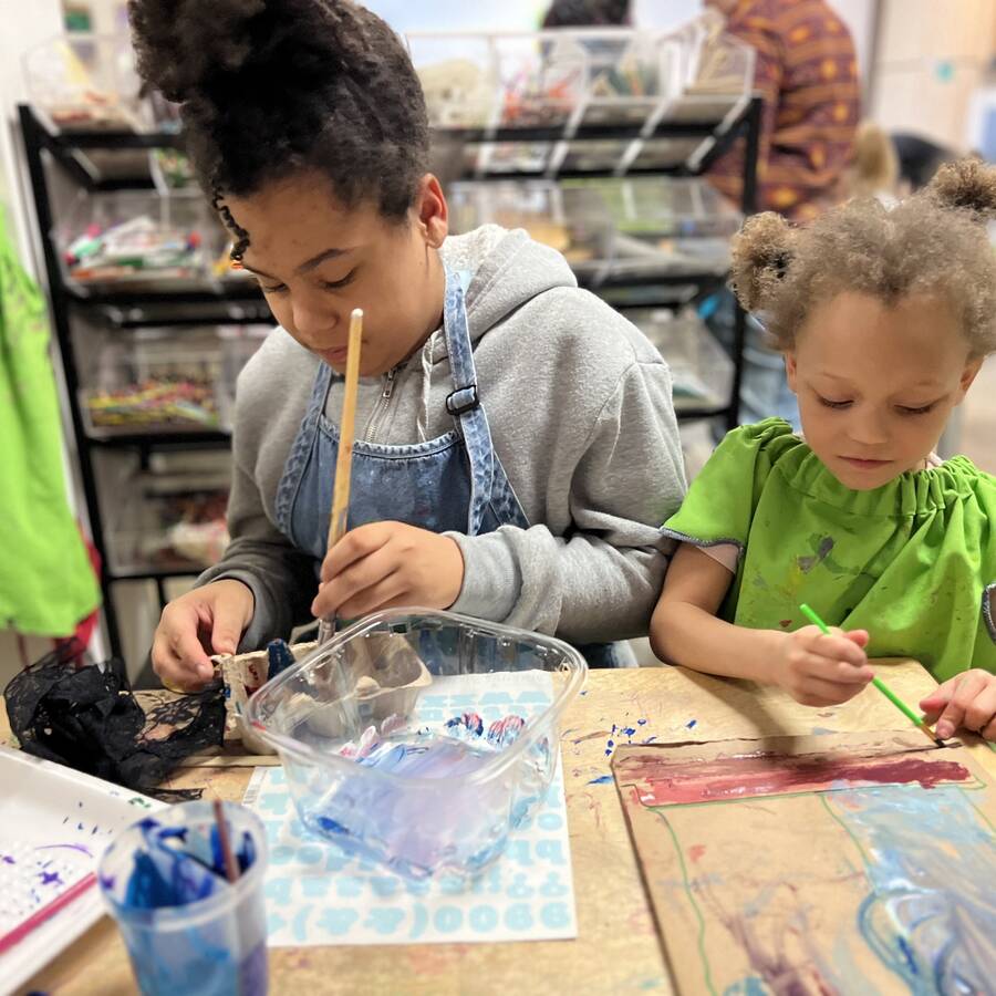 Two children sitting at a table painting on paper and cardboard.
