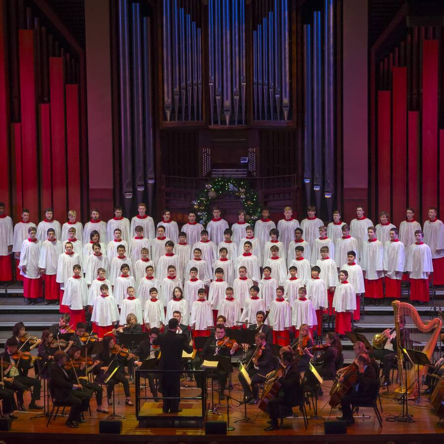 Group of boys performing on stage in white robes with an orchestra in the foreground.
